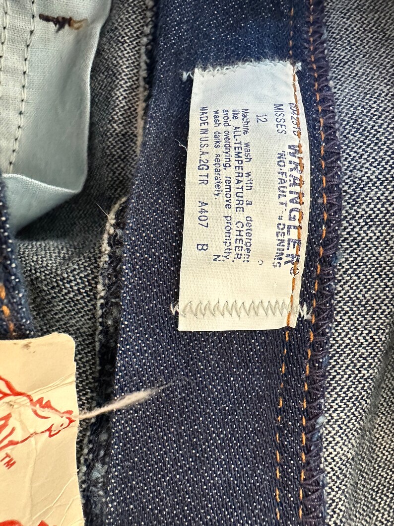 K&ouml;nnte beinhalten: Nahaufnahme einer dunkelblauen Jeans mit einem Etikett mit der Aufschrift "Wrangler No-Fault Denim". Das Etikett enth&auml;lt auch Waschanweisungen und den Text "Made in U.S.A.2G TR". Die Jeans hat Gr&ouml;&szlig;e 12.