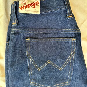K&ouml;nnte beinhalten: Dunkelblaue Jeans aus Denim mit klassischem F&uuml;nf-Taschen-Design. Die Ges&auml;&szlig;tasche hat eine charakteristische W-f&ouml;rmige Naht in einem kontrastierenden gelben Faden. Ein Etikett mit dem Wrangler-Logo ist am Bund befestigt.
