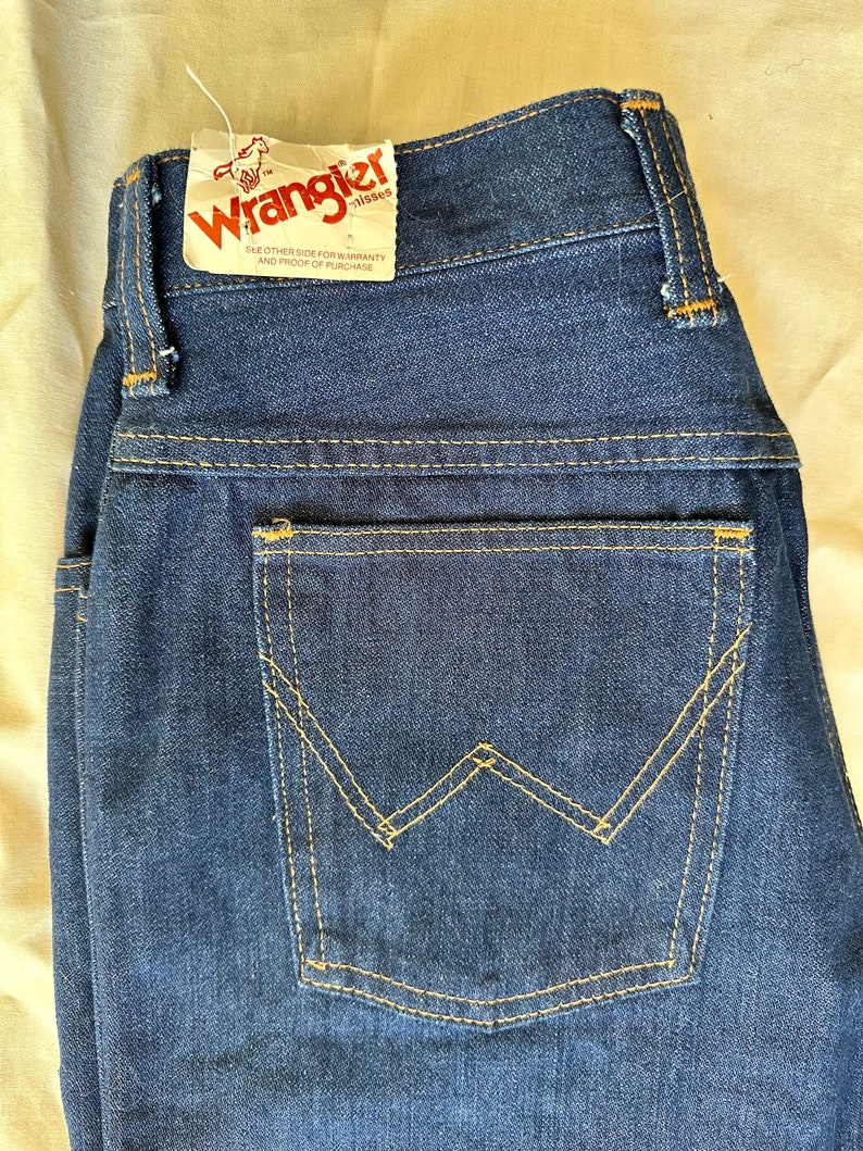 K&ouml;nnte beinhalten: Dunkelblaue Jeans aus Denim mit klassischem F&uuml;nf-Taschen-Design. Die Ges&auml;&szlig;tasche hat eine charakteristische W-f&ouml;rmige Naht in einem kontrastierenden gelben Faden. Ein Etikett mit dem Wrangler-Logo ist am Bund befestigt.
