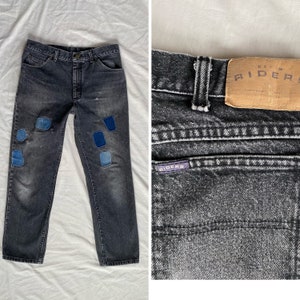 Op de afbeelding: Zwarte denim jeans met blauwe denim patches en een bruin leren patch met de tekst "DENIM RIDERS" op de achterzak.