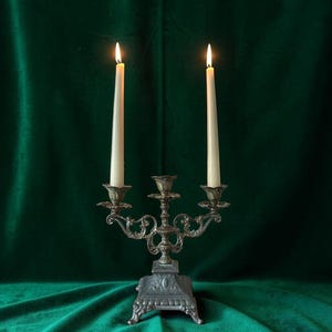 Può includere: Un candelabro in metallo con una finitura scura e ornata, dotato di tre portacandele. Due candele affusolate color bianco sporco sono accese, con fiamme visibili in cima. La base quadrata e decorativa sostiene bracci curvi.
