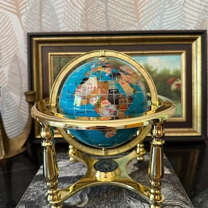 Könnte beinhalten: Ein dekorativer Globus mit einer leuchtend blauen Meeresoberfläche und Kontinenten aus eingelegten, bunten Steinen. Der Globus ist in einem polierten, goldfarbenen Metallmeridianring montiert und ruht auf einem vierbeinigen Ständer mit Kompass.