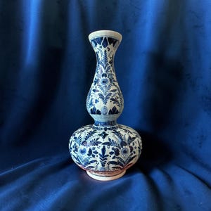 Pode incluir: Um vaso de cerâmica em forma de cabaça dupla, com uma base de cor creme decorada com intrincados padrões florais e foliares em azul escuro. O recipiente tem um pescoço estreito que se alarga no topo e um corpo largo e arredondado.