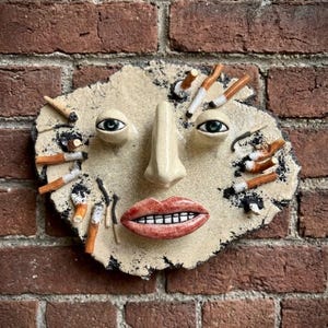 Keramisk väggmask med cigaretter, surrealistisk ansiktsskulptur, väggkonst med mörk humor