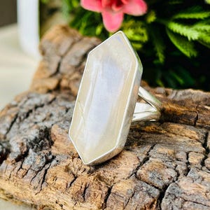 Può includere: Un anello in argento con una grande pietra bianca traslucida dal taglio esagonale allungato, incastonata in una montatura in metallo liscio. La pietra ha un aspetto lattiginoso e opalescente con sottili inclusioni su una fascia in argento.