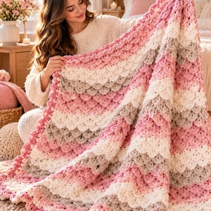Könnte beinhalten: Eine handgefertigte Häkeldecke mit einem muschelförmigen Wellenmuster in einem Farbverlauf aus zartem Rosa, Creme und Hellgrau. Das Textil ist mit einem sich wiederholenden horizontalen Streifendesign konstruiert, das einen strukturierten Effekt erzeugt.