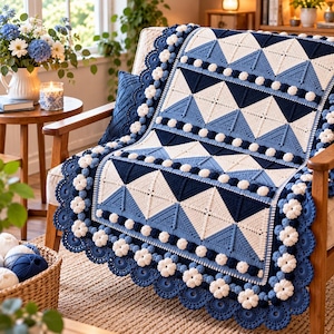 May include: A crochet blanket featuring a geometric pattern of navy blue, light blue, and white triangles. The blanket is bordered by a decorative trim of white crocheted flowers and small navy blue bobbles, creating a textured edge.