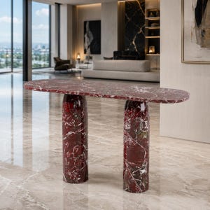 May include: A console table crafted from deep red marble with prominent white veining. The piece features a long, thin, oval-shaped tabletop supported by two thick, cylindrical columns that extend to the floor, creating a solid structure.