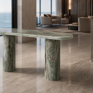 May include: A console table crafted from green marble featuring an elongated oval top with a smooth surface. The table is supported by two cylindrical, vertically fluted columns that match the green marble of the tabletop.