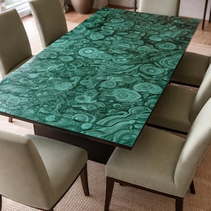 Op de afbeelding: Een rechthoekige eettafel met een levendig groen malachietpatroon met concentrische cirkelvormige motieven, ondersteund door een donkere basis, omringd door zes gestoffeerde stoelen met lichtbeige stof en donkerhouten poten.