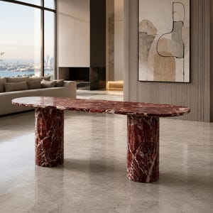 May include: A long, oval-shaped dining table crafted from deep red marble with prominent white veining. The table features a smooth, polished surface supported by two thick, cylindrical marble pedestals in the same matching red stone.