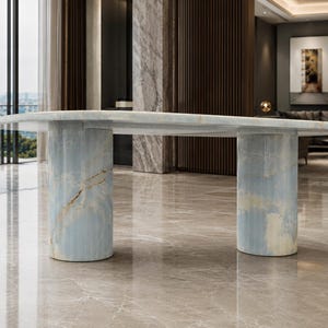 Puede incluir: Una mesa de comedor con un tablero ovalado liso de piedra azul claro con vetas blancas y doradas. La mesa se apoya en dos pedestales cilíndricos gruesos que combinan con el tablero, mostrando un patrón de mármol azul consistente.