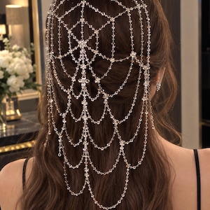 Op de afbeelding: Een zilverkleurig hoofdaccessoire met een fijn webachtig patroon van onderling verbonden kettinkjes en kleine heldere kristallen. Het sieraad valt over de achterkant van het hoofd, waarbij meerdere lagen een trapsgewijs effect creëren.