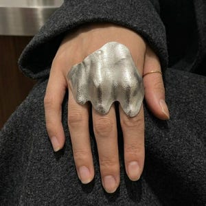 May include: A large, silver-toned statement ring featuring an irregular, wavy, and textured surface that spans across three fingers. The metal has a matte, brushed finish with organic, undulating contours that create a sculptural, layered appearance.