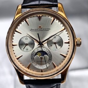 Può includere: Un orologio da polso Jaeger-LeCoultre con cassa in oro rosa, quadrante argentato con finitura soleil e cinturino in pelle nera. Il quadrante presenta due sotto-quadranti, un indicatore delle fasi lunari e la data. JAEGER-LECOULTRE e AUTOMATIQUE sono visibili.