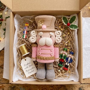 Puede incluir: Un muñeco cascanueces de ganchillo con sombrero beige, chaqueta rosa y botas grises, en una caja con un carrete de hilo dorado, una etiqueta que dice Crafted with Love-Nutcracker Crochet, dos pequeños adornos de cascanueces y acebo.