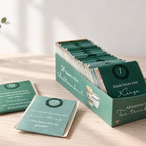 May include: A rectangular green box containing a set of cards, with two additional cards placed in front. The box and cards feature the German text 24 Tassen voller Achtsamkeit, Zünde heute eine Kerze, Abwarten und Tee trinken, and numbered circles.