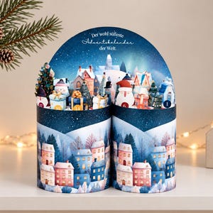 May include: A cylindrical cardboard advent calendar featuring a winter village scene with blue and pink houses, snow-covered trees, and gift boxes. The top arch displays the German text 'Der wohl süßeste Adventskalender der Welt' above numbered compartments.