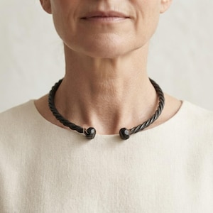 Könnte beinhalten: Eine dunkelbraune, gedrehte Metall-Torque-Halskette mit starrem, offenem Design. Das Band hat eine spiralförmige, seilartige Textur, die zu zwei glatten, abgerundeten, knollenartigen Endstücken an der Vorderseite führt, die am Halsansatz liegen.