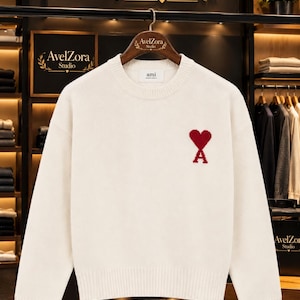 Puede incluir: Un suéter de punto de manga larga en color crema con cuello redondo y un logotipo rojo en forma de corazón con la letra A debajo en el pecho izquierdo. La etiqueta interior del cuello muestra el texto ami.