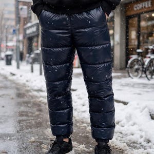 May include: Dark navy blue quilted puffer trousers with a shiny, water-resistant finish. These insulated trousers feature horizontal stitching, an elasticated waistband, and cuffed ankles, paired with black athletic trainers.