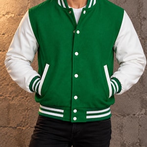 Puede incluir: Una chaqueta universitaria con cuerpo de mezcla de lana verde y mangas blancas. La chaqueta presenta cuello, puños y dobladillo acanalados con rayas blancas, además de cierres de botones a presión blancos y dos bolsillos laterales con ribete blanco.