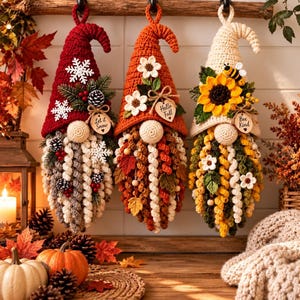 May include: Three hanging crochet gnomes with pointed hats. The left gnome is red with snowflakes and a tag reading Let it Snow. The center gnome is orange with flowers and a tag reading Hello Fall. The right gnome is cream with a sunflower, bee, and a tag reading Bee Kind.