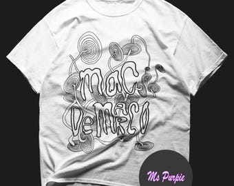 Op de afbeelding: Een wit t-shirt met korte mouwen voorzien van een zwarte lijntekening van meerdere paddenstoelachtige vormen en de tekst "mac de marco" in het midden. Een donker rond embleem in de rechterbenedenhoek toont de tekst "Ms Purpie" in het roze.