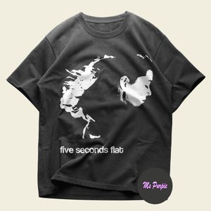 Puede incluir: Una camiseta de manga corta gris carbón con un estampado gráfico blanco de la silueta de perfil de una persona y el texto five seconds flat. El diseño está situado en la zona del pecho de la prenda, que tiene un cuello redondo.