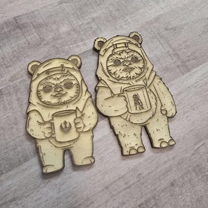 May include: Two wooden cutouts of Ewoks, each holding a mug. The Ewoks are light brown with detailed facial features and are wearing hooded outfits. The mugs have a logo on them. The cutouts are placed on a light gray wooden surface.