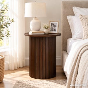 May include: A cylindrical side table featuring a dark brown wood finish with vertical slats along the base and a smooth circular top. The table supports a white ceramic lamp, a stack of books, a small vase with white flowers, and a picture frame.
