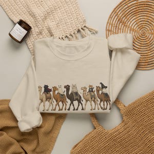 May include: A cream-colored crewneck sweatshirt featuring a graphic print of six camels walking in a line. Each camel carries a cat dressed in a different costume, including a ghost, a mummy, a skeleton, and a witch, all rendered in a detailed illustration.