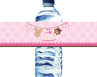 May include: Pink and brown baby shower water bottle label with a polka dot background. The label features a teddy bear, a bib, and two baby outfits hanging on a clothesline.