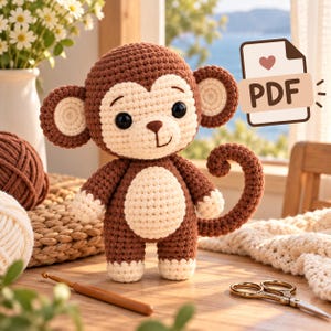 Op de afbeelding: Een gehaakt amigurumi-aapje in bruin en crème met grote zwarte ogen, een gekrulde staart en een lichte buik. Een documentpictogram met de tekst PDF en een hartsymbool bevindt zich in de rechterbovenhoek van de afbeelding.