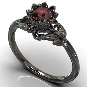 Pode incluir: Um anel de metal preto com uma pedra preciosa vermelho-escura de corte redondo ao centro. O engaste tem a forma de uma flor a desabrochar com pétalas a rodear a pedra, e a banda apresenta motivos esculpidos de folhas e videiras.