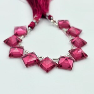 May include: A strand of faceted, square-shaped, deep pink gemstones arranged in a curved line. Each gemstone is separated by small, cylindrical silver-toned spacers. A dark pink tassel is attached to the top of the gemstone strand.