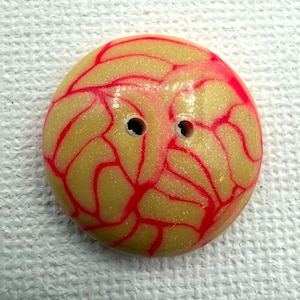 May include: A round, two-hole button featuring a pale, glittery yellow base with a vibrant, raised red vein-like pattern across the surface. The button has a smooth, glossy finish and a slightly domed shape, providing a distinct contrast between the colors.