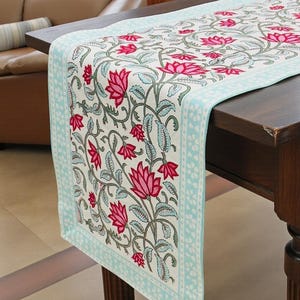 May include: A rectangular table runner featuring a cream-coloured base with a repeating pattern of vibrant pink lotus flowers and green leafy vines. The runner is bordered by a light blue band with a white floral motif and a small logo in the corner.