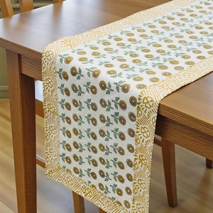 May include: A rectangular table runner featuring a white base with a repeating pattern of small yellow flowers and green leaves. The runner is bordered by a wide yellow and white geometric print. A small logo in the corner reads Sanwariya Living Co.