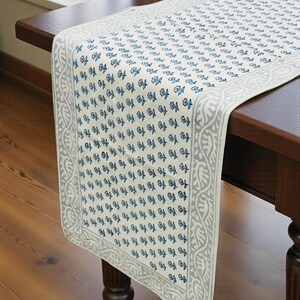 May include: A rectangular table runner featuring a repeating pattern of small blue and grey floral motifs on an off-white background. The runner is finished with a wide, decorative grey border displaying a continuous organic leaf-like design.