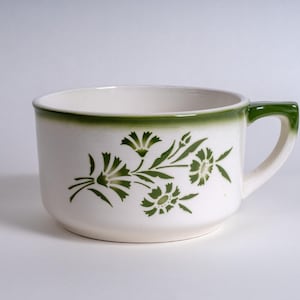 Op de afbeelding: Een witte keramische soepkom met een groen bloemmotief aan de zijkant en een groene rand. De kom heeft één handvat aan de rechterkant, met een groen accent op de bovenkant van het handvat. Het ontwerp bestaat uit gestileerde groene bloemen.