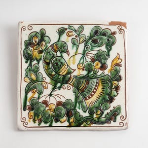 Op de afbeelding: Een vierkante keramische tegel met een handgeschilderd ontwerp van een vogel omringd door gestileerd groen en geel gebladerte. Het kunstwerk gebruikt een palet van donkergroene, gele en bruine glazuren op een crèmekleurige achtergrond met rand.