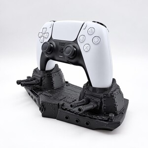 May include: A black battleship-themed controller stand featuring two turret-style cradles, holding a white and black wireless game controller with dual analogue sticks, a directional pad, and four action buttons marked with geometric symbols.