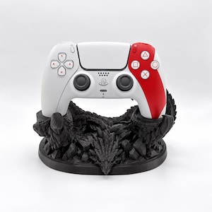 May include: A white and red wireless video game controller resting on a black, textured dragon-themed display stand. The controller features a central PlayStation logo, two black analog sticks, a directional pad, and four action buttons with red outlines.