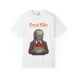 May include: A white short-sleeved t-shirt featuring a graphic of a skeletal figure in a brown plaid blazer and red polka-dot tie holding a bowl of cereal. The text Cereal Killer and Crypt Delights For Your Demise is printed above the figure.