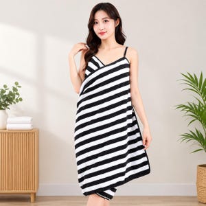 May include: A wearable towel wrap featuring a black and white horizontal striped pattern. The garment has thin shoulder straps and a wrap-around design that secures at the front, crafted from a soft, absorbent fabric with a textured finish.