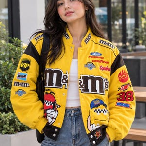 May include: A bright yellow and black varsity-style jacket featuring M&M's branding, NASCAR logos, and various sponsor patches including Goodyear, Combos, and Master Tools. The jacket displays a large M&M's logo and cartoon character graphics.