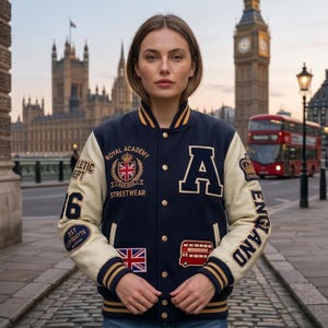 Peut inclure: Une veste universitaire bleu marine avec des manches crème, ornée d'écussons incluant le texte Royal Academy, Athletic Dept, England, un drapeau Union Jack, un bus rouge à impériale, une couronne et la lettre A sur la poitrine.