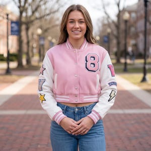 May include: A light pink and white varsity jacket featuring a snap-button front, striped ribbed cuffs, and a collar. The jacket displays a large number 8 on the chest, with various colourful patches including stars, a lightning bolt, and text on the sleeves.