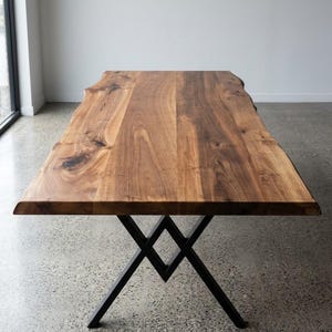 Peut inclure: Une table à manger rectangulaire dotée d'un plateau en bois massif à bord naturel avec des motifs de grain et des nœuds visibles. La table est soutenue par une base en métal noir composée de deux cadres en X entrecroisés formant un losange central.